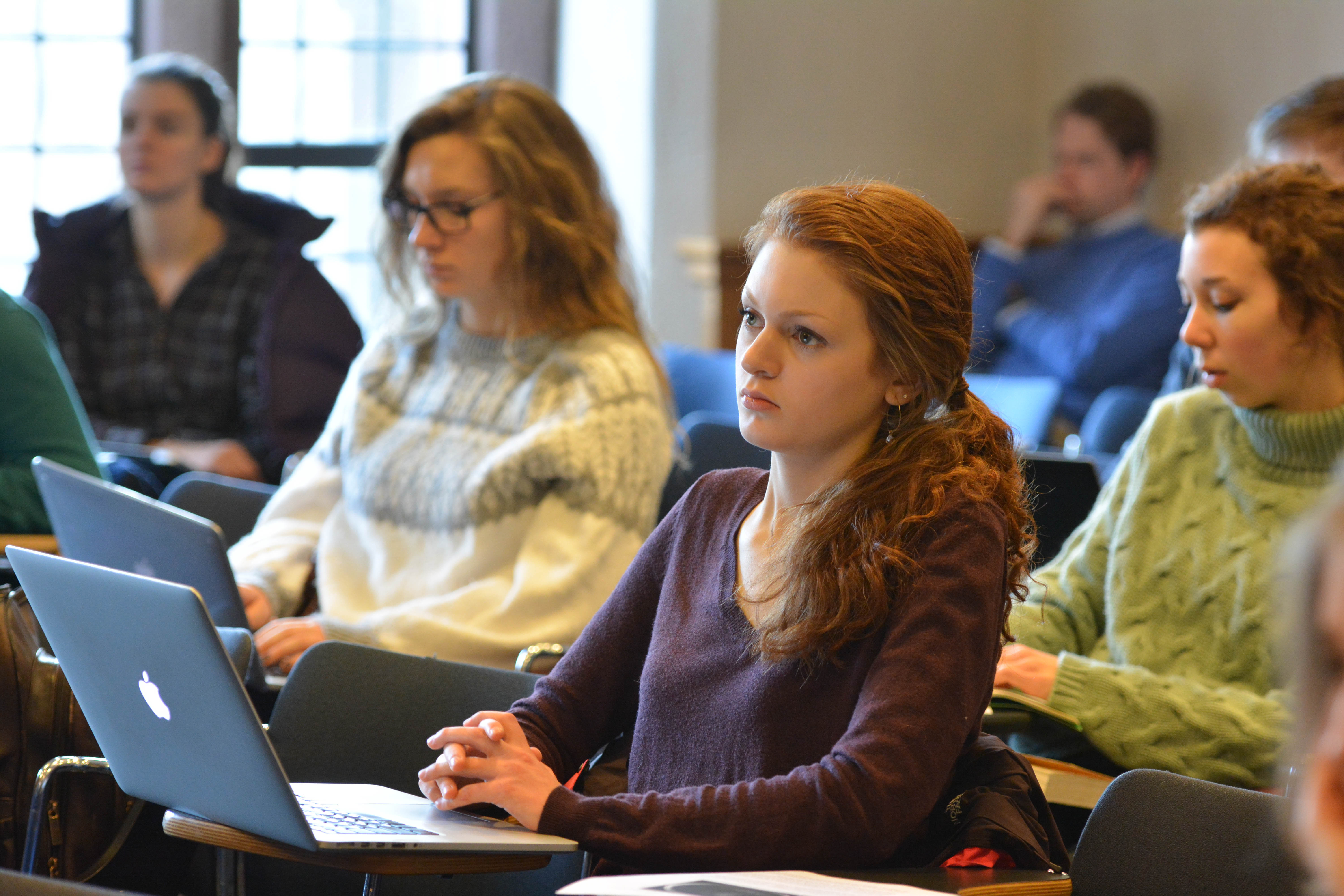 A college student at a table reviewing printed course syllabi next to a laptop