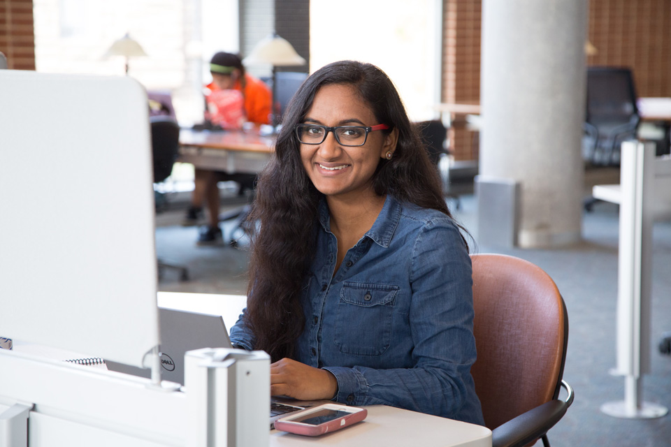 A college student wearing a campus ID badge working at a library circulation desk, assisting a patron at the counter