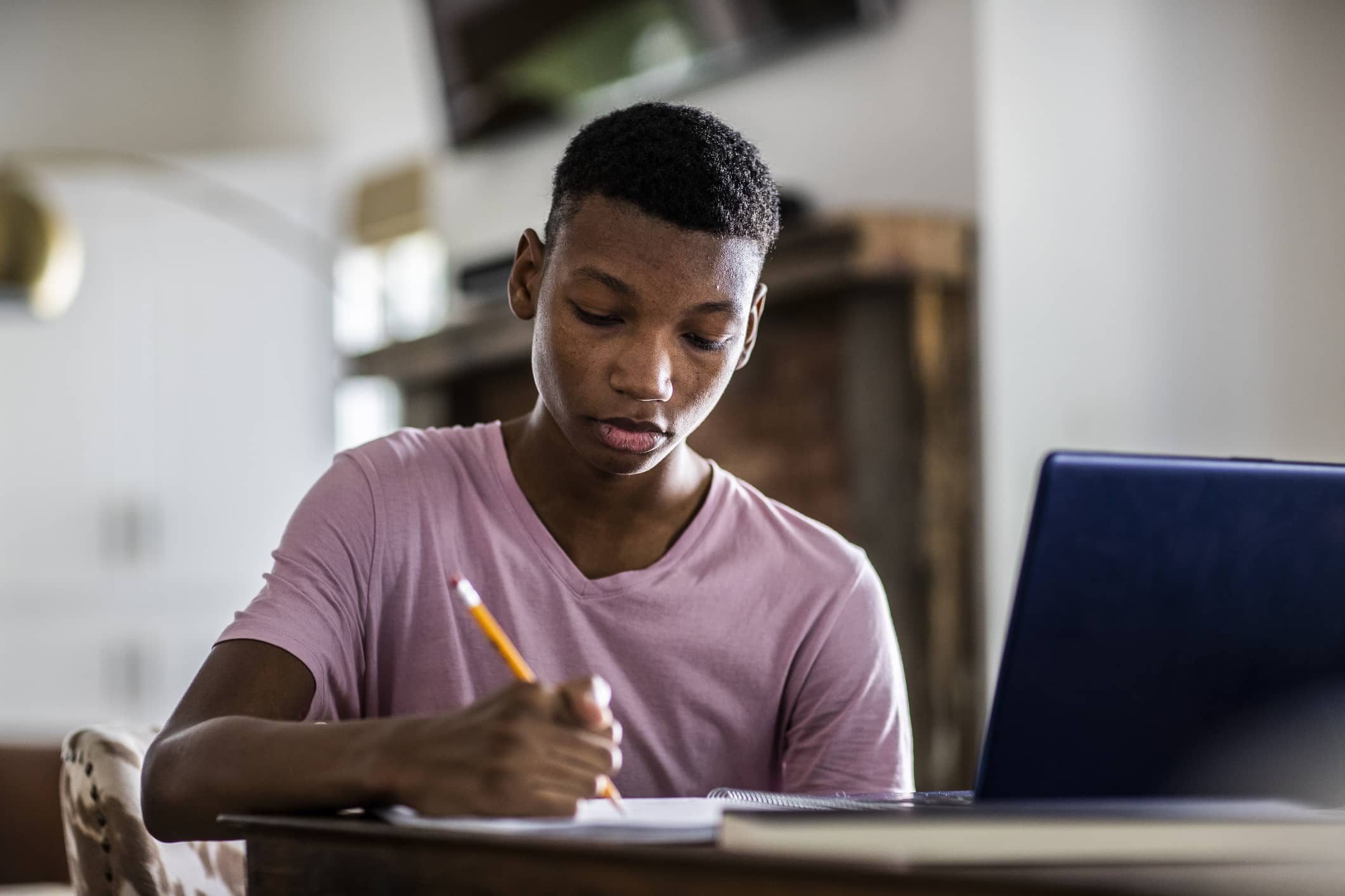 A high school student sitting at a desk at home, laptop open with a notebook and pen nearby, pausing thoughtfully while starting a college application essay