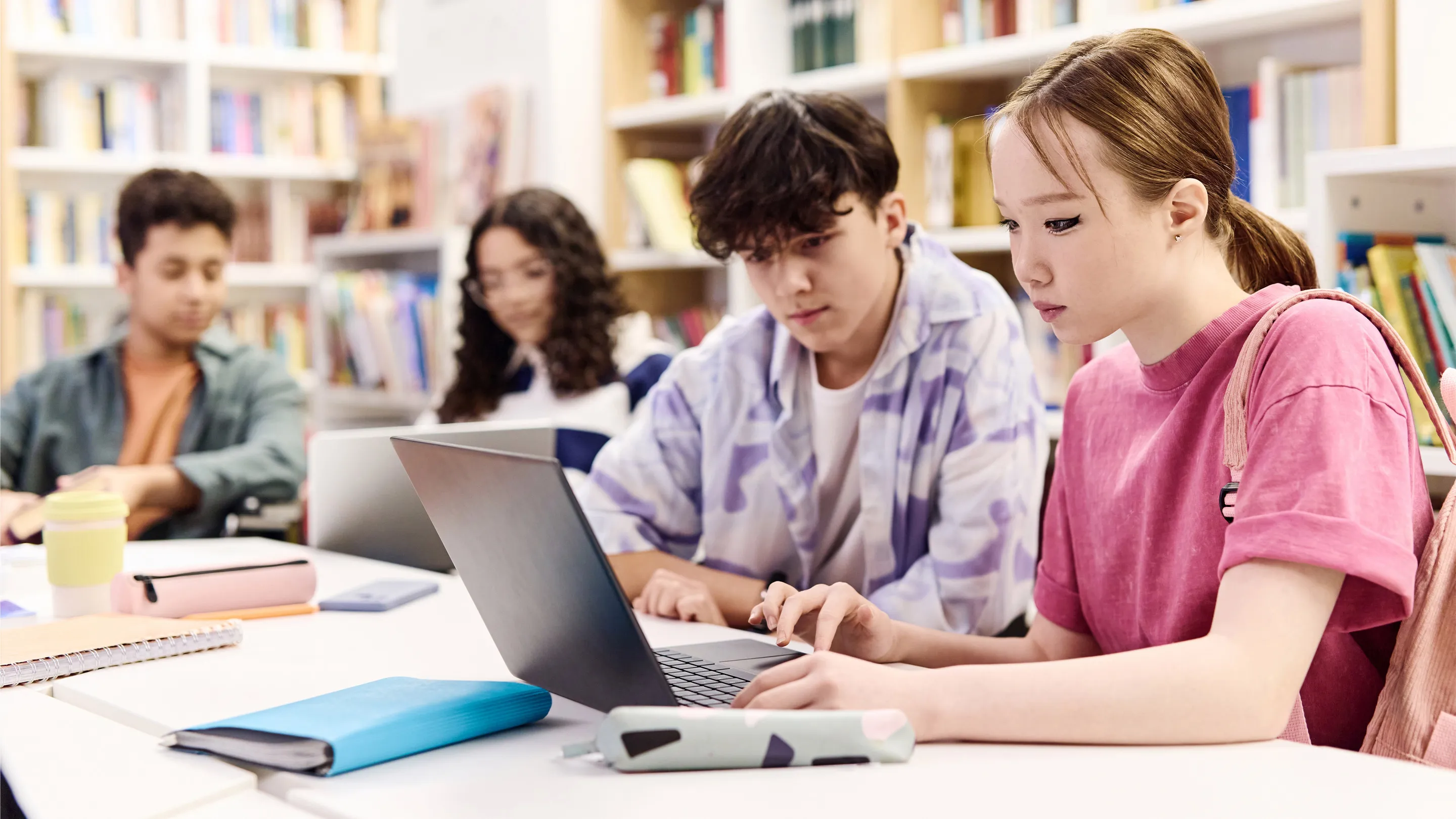 A high school student studying at a library table with notebooks and a laptop, softly lit, candid school day moment