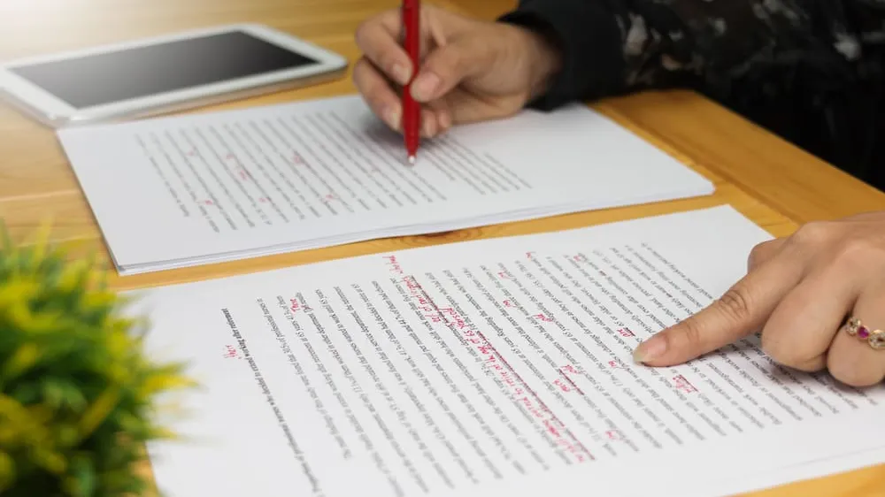 A student sitting at a desk marking edits on a printed essay with a pen, with a laptop closed nearby