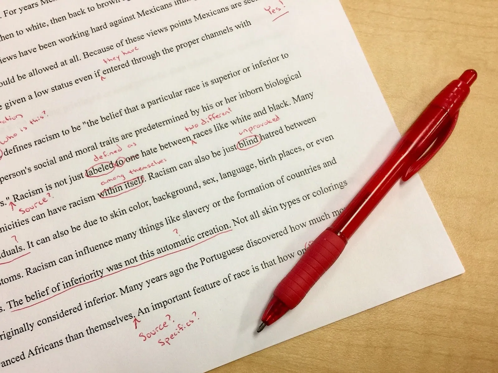 A student sitting at a desk reviewing a printed essay and making edits with a red pen next to an open laptop