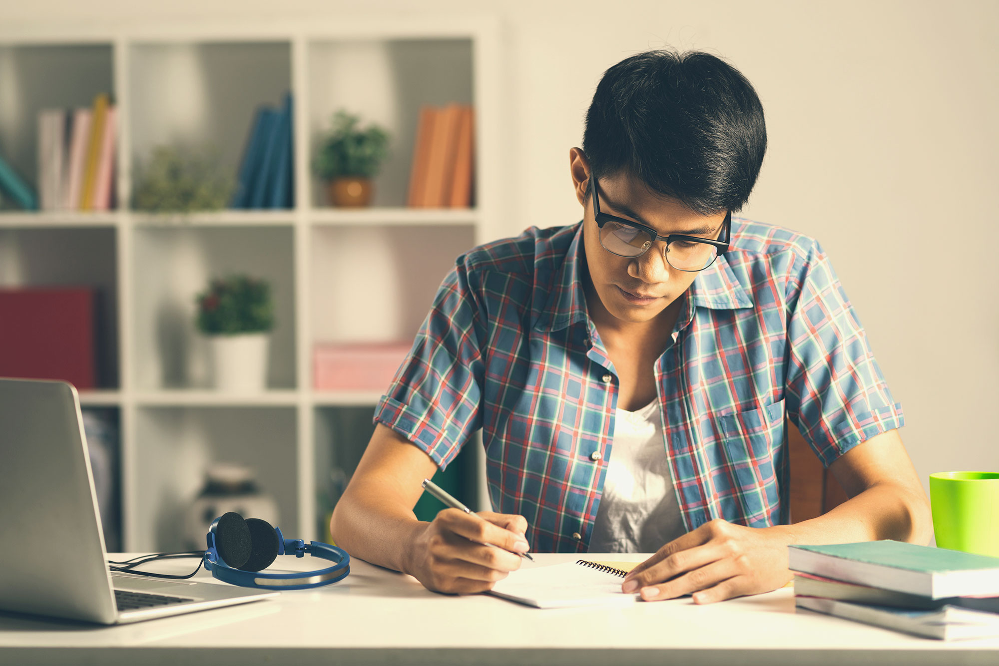 A student sitting on a bed with a laptop open and printed pages beside them, marking revisions on a college essay draft with a pen