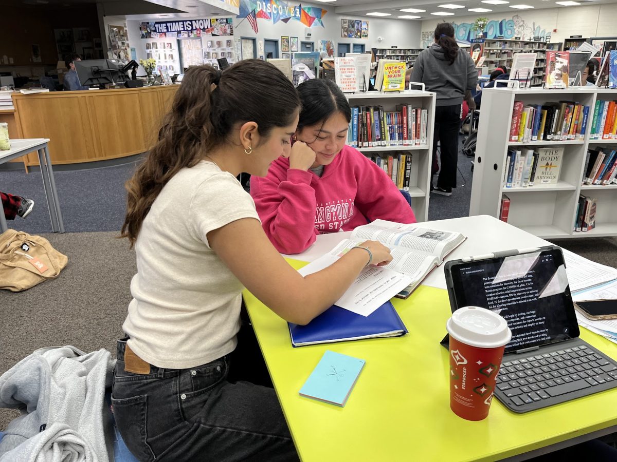 A student tutor sitting with a younger student at a school library table, pointing at a worksheet while both focus on the work