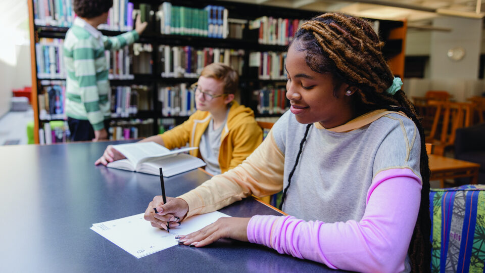 A student typing on a laptop in a quiet library with a notebook beside the computer, focused on drafting an essay
