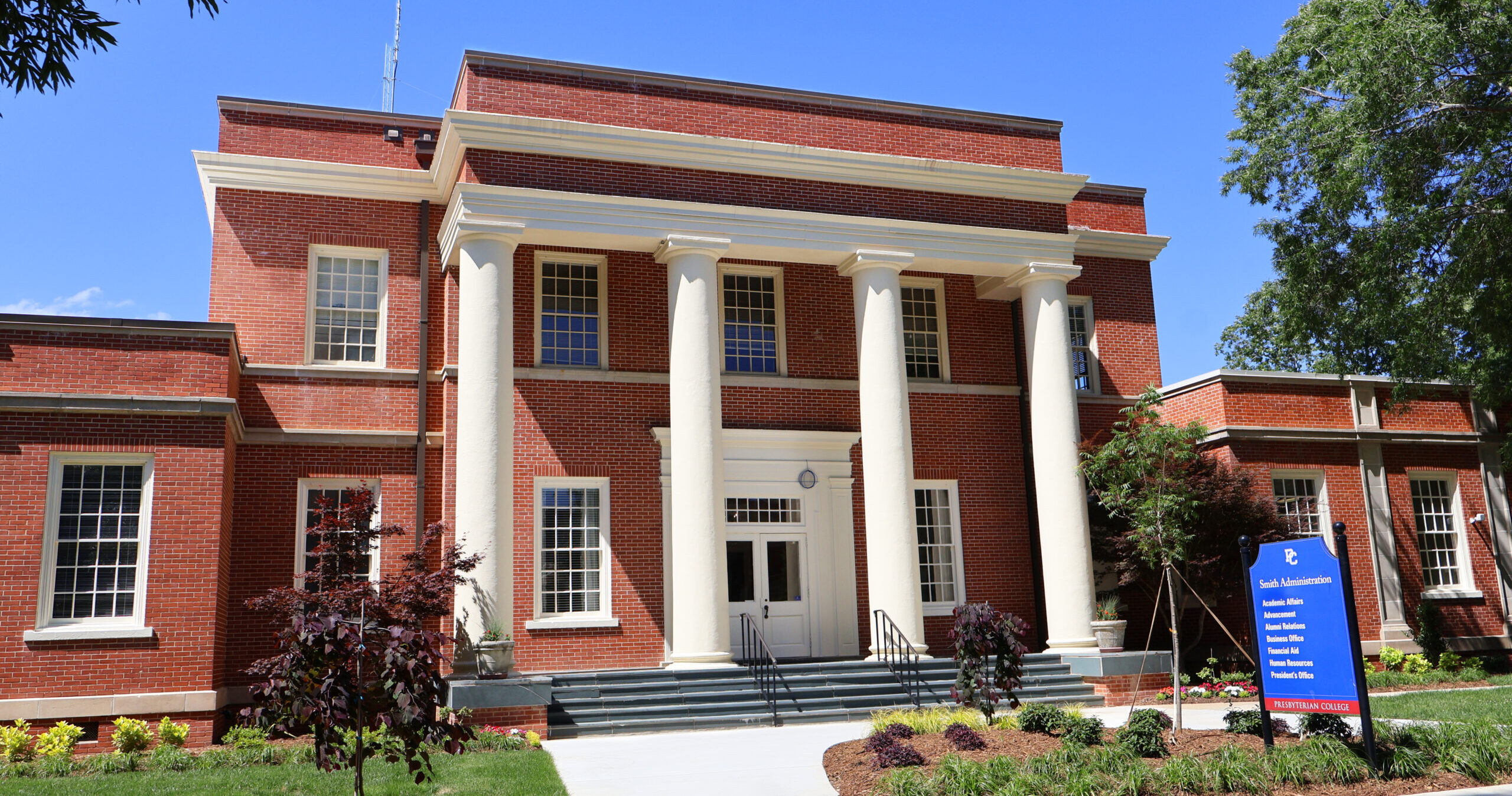 A student walking toward the entrance of a university administrative building on campus in daylight, carrying a backpack and holding paperwork