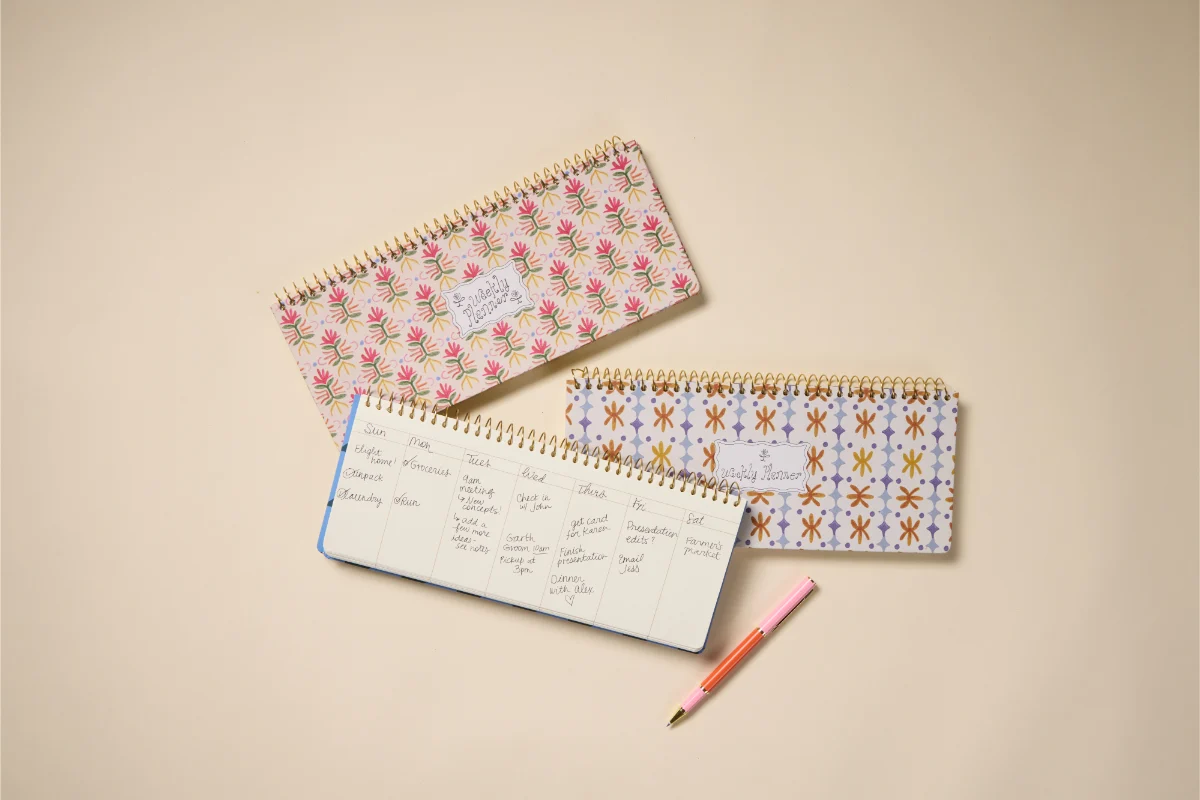An open paper planner on a desk with colored pens and a few children’s books nearby, showing a calm planning setup